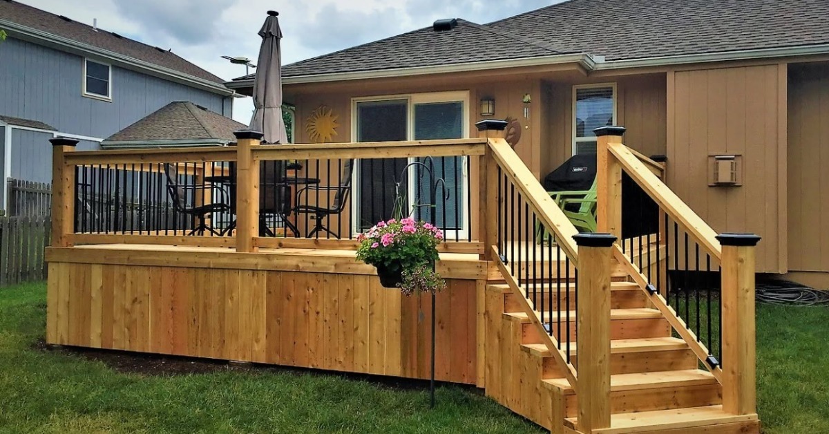A wooden backyard deck with stairs, black railings, patio furniture, a grill, and a hanging flower basket.
