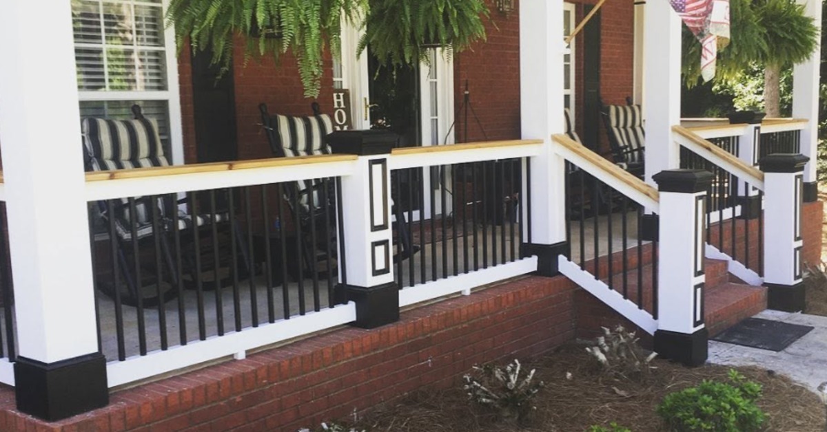 A covered brick porch with white columns, black railings, striped chairs, hanging ferns, and an American flag.