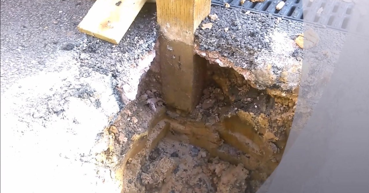 A close-up of a hole carved out on a concrete floor, with a wooden beam digging into the ground next to a metal drain. A close-up of a hole carved out on a concrete floor, with a wooden beam digging into the ground next to a metal drain.