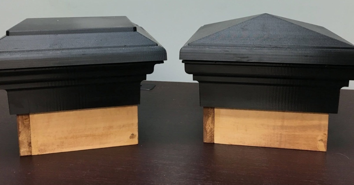 A close-up of two black post caps attached to short wooden blocks, resting on a dark brown wooden table surface. A close-up of two black post caps attached to short wooden blocks, resting on a dark brown wooden table surface.