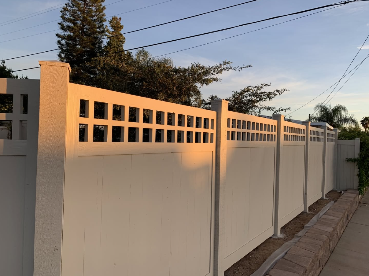 The top of a white wooden fence is illuminated by soft light coming from the sunset, with trees visible in the background. The top of a white wooden fence is illuminated by soft light coming from the sunset, with trees visible in the background.