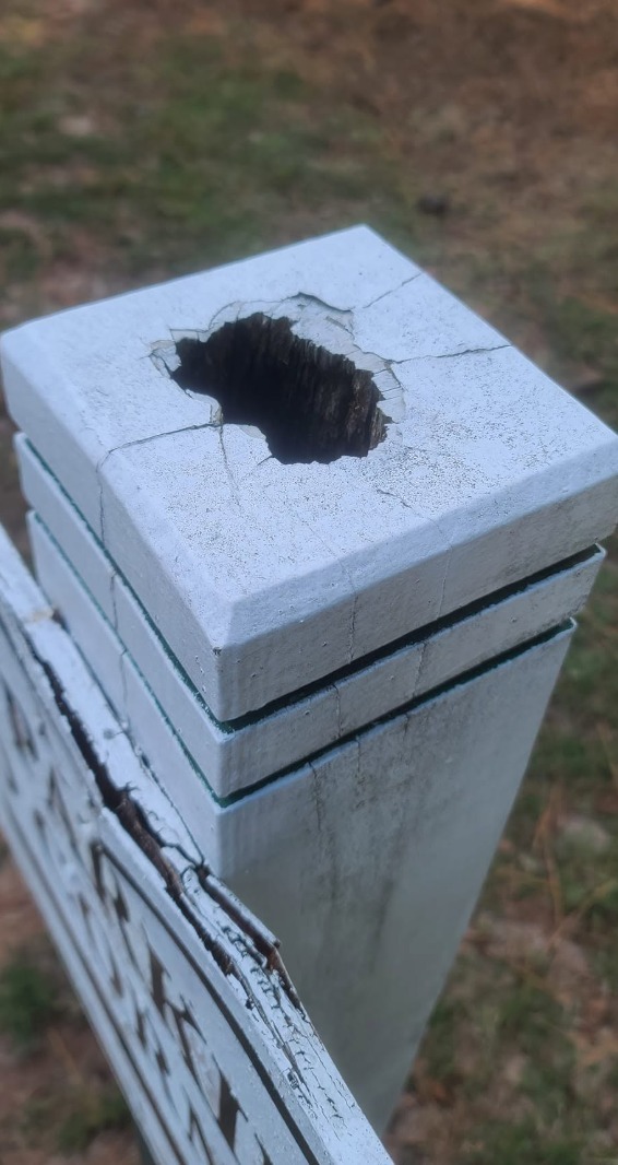 A close-up of a white post cap with a large dark hole, cracked edges, and chipped paint above lattice.