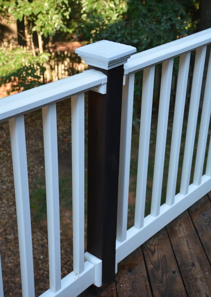 A black deck post with a white cap and white railings attached to a wooden floor. Trees are visible in the background.