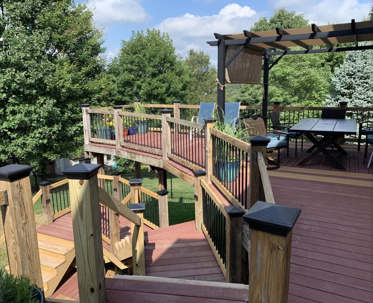 How To Measure Your Deck or Fence for Post Caps A large raised deck with wood railings, black balusters, stairs, patio furniture, and trees surrounding it.
