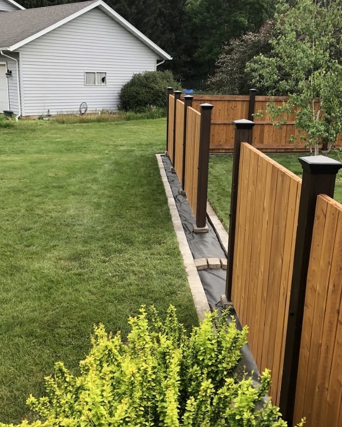 A close-up of a wooden privacy fence with dark posts along a green yard near shrubs, trees, and a white house.