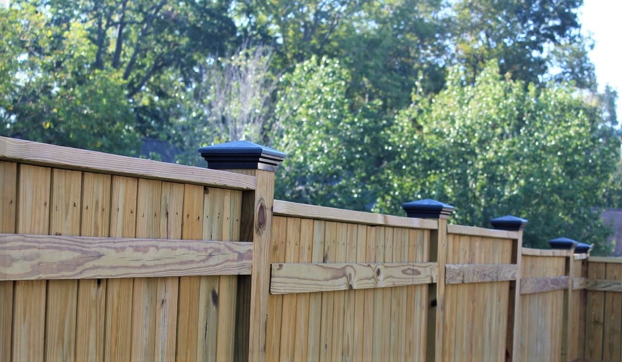What Are the Benefits of HDPE for Post Caps? A wooden privacy fence with black post caps extends along a yard with trees and greenery in the background.