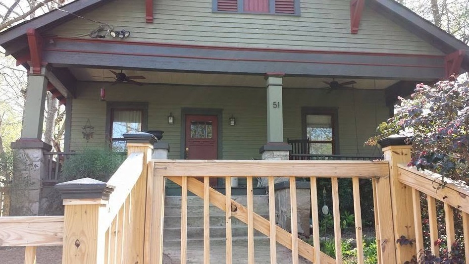 A wooden stair railing leading toward the covered front porch of a green house with red trim and a central door.